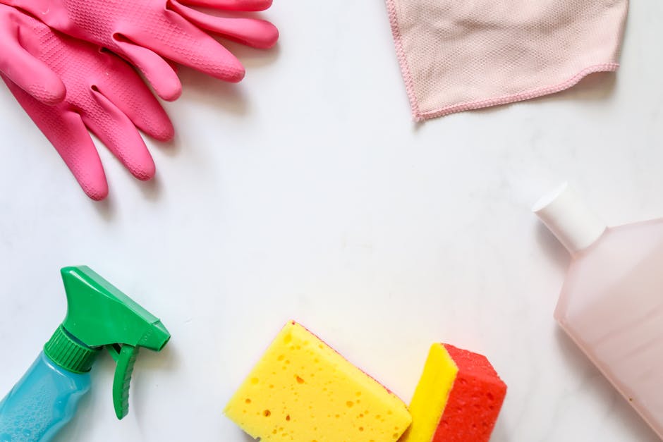 Arrangement of cleaning tools and supplies on a white surface, including a pair of pink rubber gloves, a pink cloth, a green spray bottle, a yellow and red sponge, and a white bottle of cleaning solution, illustrating the process of surface cleaning and hygiene maintained by Harrow Cleaners in their end of tenancy cleaning services offered at harrow-cleaners.co.uk.