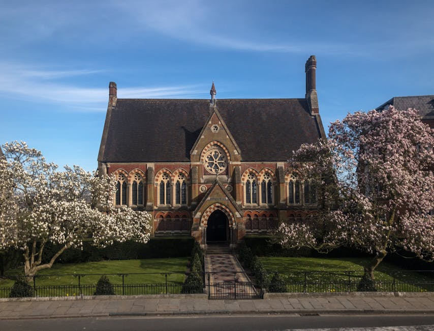 The exterior of a historic church building with a steep, dark shingle roof, brick and stone walls featuring Gothic-style arched windows, and decorative stone detailing around the entrance. The church is surrounded by blooming trees with white and pink flowers, neatly maintained green lawns, and a black metal fence along the sidewalk under a clear blue sky, showcasing a clean and picturesque setting. Harrow Cleaners provides expert surface cleaning and sanitisation services for maintaining such architectural beauty, as highlighted in the End of Tenancy Cleaning Harrow on the Hill guide.