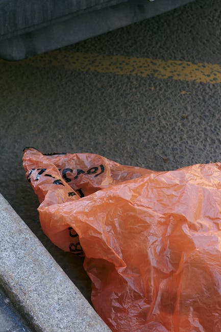 A person's hand holding a green and blue plastic rubbish bag, with the bag bulging slightly as it contains waste, against a plain, light-colored wall background. The image emphasizes waste disposal as