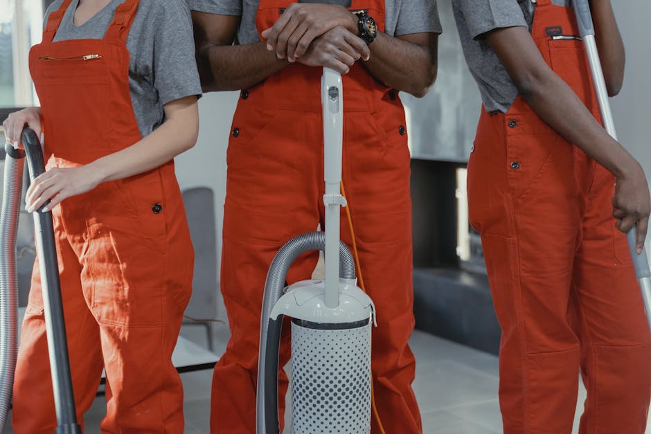 Three professional cleaners in orange overalls performing surface cleaning with vacuum cleaners in an indoor setting. The centre worker is holding a canister vacuum with a white and gray dotted design