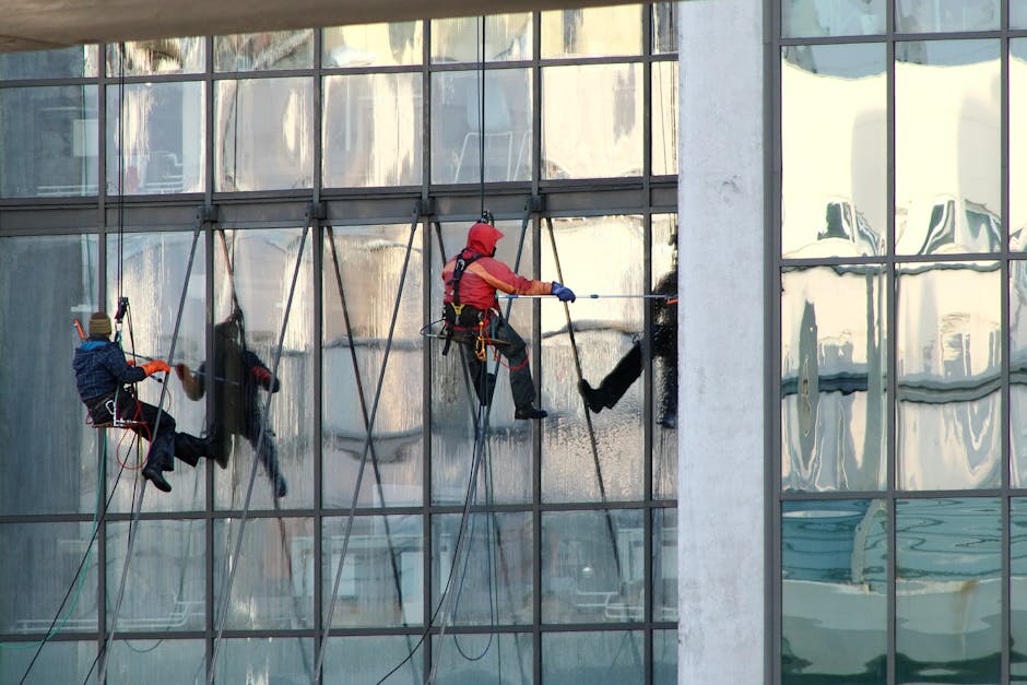 Three professional cleaners in orange overalls performing surface cleaning with vacuum cleaners in an indoor setting. The centre worker is holding a canister vacuum with a white and gray dotted design