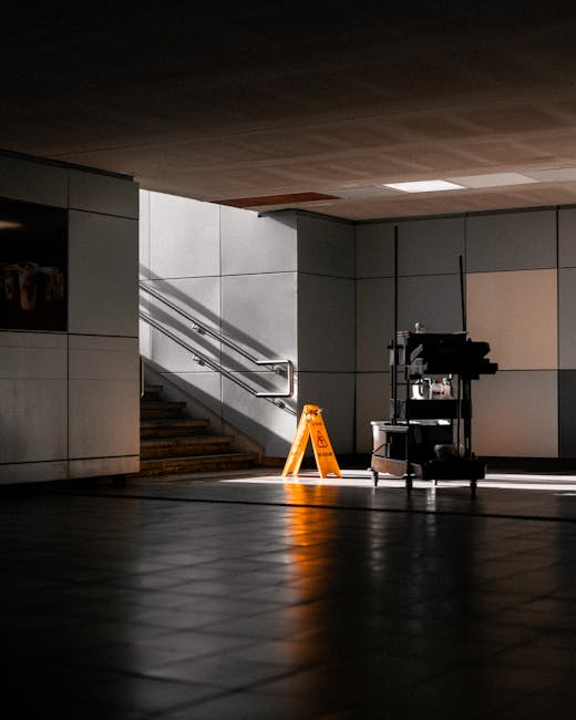 A minimalist indoor space with tiled flooring and smooth, light-colored wall panels illuminated by natural light coming from an unseen source. In the center, a cleaning cart equipped with various supp